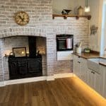 A renovated country kitchen with white cupboards, timber benchtops, and a large brick fireplace surrounding a coal range.